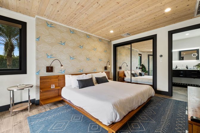 Modern coastal master bedroom with wood-paneled ceiling, king platform bed with white linens, blue-bird patterned accent wall, mirrored sliding closet, bedside reading lamps, blue geometric rug and a palm-tree view through the window.