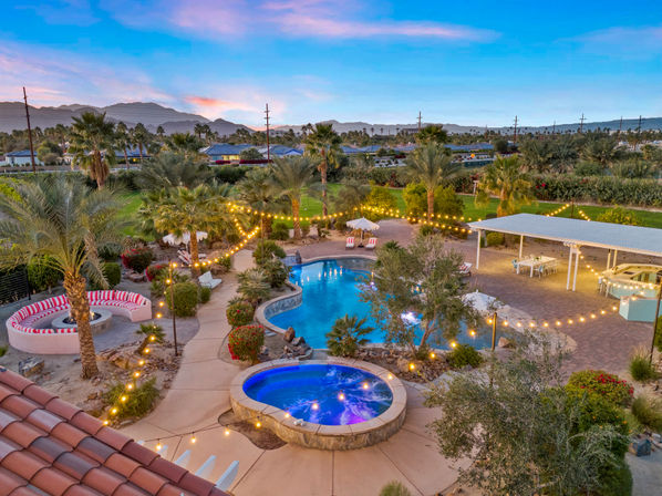 Aerial twilight view of a desert oasis backyard: glowing string lights draped over palm trees, a curving swimming pool, bubbling blue hot tub, cozy circular firepit seating, and distant mountain sunset.