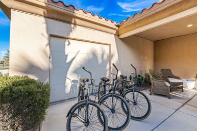 Sunlit Mediterranean-style stucco home exterior with terracotta tile roof and a palm shadow on the garage door; four black cruiser bicycles parked on the concrete driveway beside wicker patio chairs and desert plants under a bright blue sky.