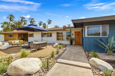 Mid-century modern Palm Springs–style desert home exterior with orange front door, blue-and-white siding, concrete walkway, outdoor seating under a large black umbrella, palm trees and drought-tolerant landscaping with boulders.