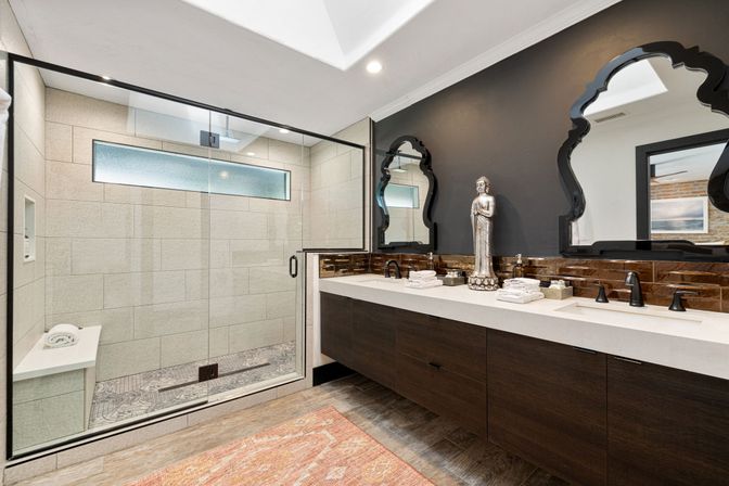 Modern spa-like bathroom with glass walk-in shower and patterned tile floor, skylight, long quartz double vanity with dark wood cabinets, ornate black-framed mirrors, bronze faucets and decorative statue.