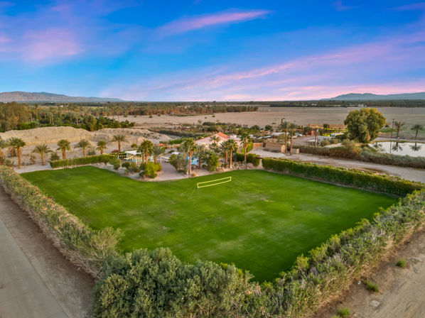 Desert oasis at sunset featuring a lush rectangular green lawn enclosed by hedges, palm trees, string lights and a backyard volleyball net, with arid fields and distant mountains under a pink-purple sky.