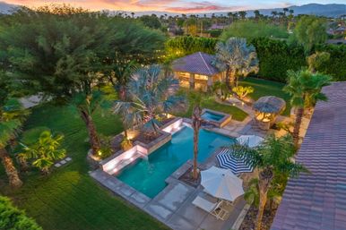 Aerial view of a resort-style backyard desert oasis at sunset with an illuminated rectangular pool and raised spa, palm trees, tiled-roof cabana, thatched palapa, striped umbrella and lounge chairs on a concrete patio with distant mountains.