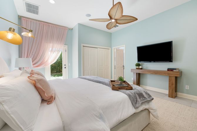 Bright sunlit modern bedroom with king bed in white linens, pastel blue walls, pink sheer curtains over a glass door, wooden leaf-style ceiling fan, wall-mounted TV and rustic wood console with potted plants — cozy coastal-inspired retreat.