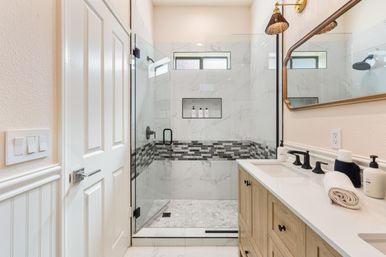 Bright modern bathroom with frameless glass shower, marble-look wall tiles and gray mosaic accent band, built-in shower niche with bottles, wood vanity with white countertop, black matte faucets and rolled towels.
