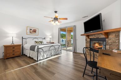 Bright cozy bedroom with iron bed, hardwood floors, ceiling fan, stone fireplace with mounted TV, wooden desk and sliding glass door to sunny patio.