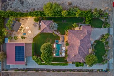 Aerial drone view of a sunlit suburban estate with terracotta-tiled roof, rectangular pool and spa, solar panels on garage, string-lit patio, palm trees and manicured lawn.