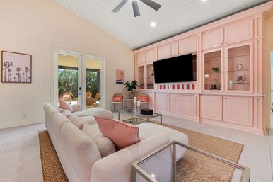 Bright coastal-inspired living room with plush white sectional and coral pillows, pastel pink built-in cabinets with wall TV, glass coffee table on woven rug, ceiling fan and French doors opening to a sunny patio.