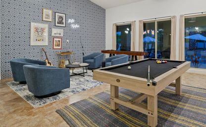 Bright residential game room with light-wood pool table (black felt) and shuffleboard, four blue lounge chairs around a round coffee table, patterned blue accent wall with framed art and a guitar in the corner, plus large windows overlooking a patio.