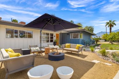 Sunny mid-century modern patio with a central fire pit, large black umbrella, outdoor sofas with yellow pillows, palm trees and mountain backdrop