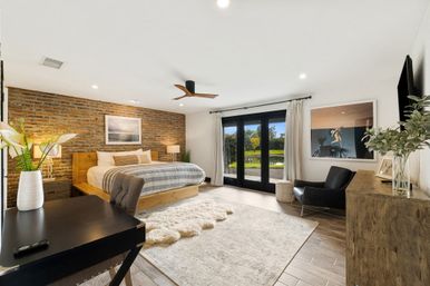 Sunlit modern rustic master bedroom with exposed brick accent wall, wooden platform bed, striped bedding, shag rug, desk and lounge chair, and black patio doors opening to a pond and palm-lined yard.