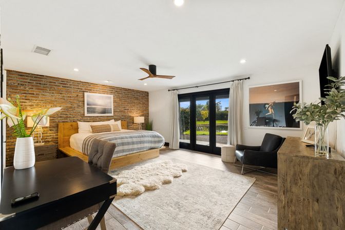 Sunlit modern rustic master bedroom with exposed brick accent wall, wooden platform bed, striped bedding, shag rug, desk and lounge chair, and black patio doors opening to a pond and palm-lined yard.