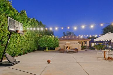Evening backyard patio with a portable basketball hoop and ball on a concrete court, string lights overhead, lush hedges, outdoor seating around a fireplace, and a tiled‑roof pavilion.