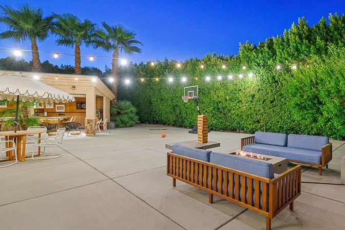 Evening residential backyard patio for outdoor entertaining: concrete court with portable basketball hoop and ball, string lights strung under tall palm trees, covered outdoor bar and dining table with umbrella, wooden lounge sofas with blue cushions around a gas fire pit and tall privacy hedge.
