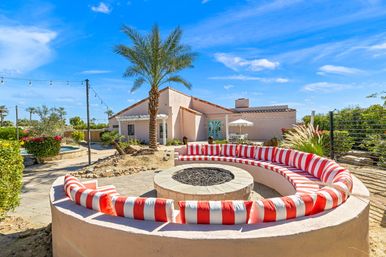 Desert backyard with curved built-in seating around a stone fire pit, red-and-white striped cushions, palm tree, stucco house and string lights under a bright blue sky.