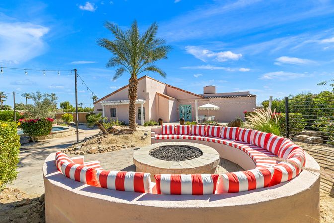Desert backyard with curved built-in seating around a stone fire pit, red-and-white striped cushions, palm tree, stucco house and string lights under a bright blue sky.