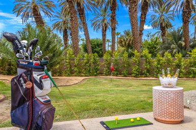 Golf bag and clubs beside a backyard putting green with yellow practice balls and a red flag, palm trees under a bright blue sky and an ice bucket of bottles on a side table.