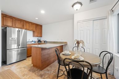 Bright modern kitchen and breakfast nook with stainless steel French-door refrigerator, warm wood cabinets, granite breakfast island, round dining table set with bowls and a vase of dried flowers, tile floor and sliding pantry doors.