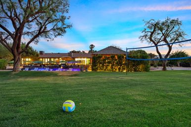 Volleyball on a green lawn in a resort-style backyard at dusk — net set up, illuminated pool, lounge chairs and umbrellas in front of a softly lit house.