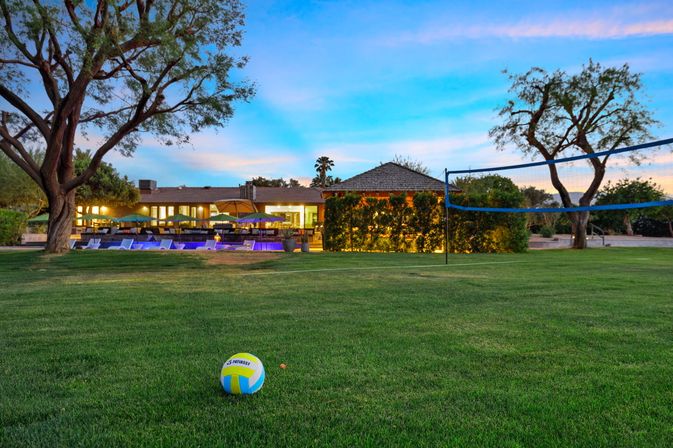 Volleyball on a green lawn in a resort-style backyard at dusk — net set up, illuminated pool, lounge chairs and umbrellas in front of a softly lit house.