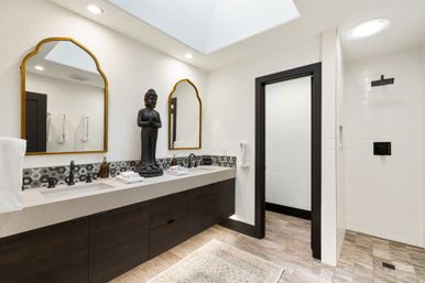 Modern spa-style bathroom with double sink vanity, dark wood cabinets and quartz countertop, two gold-framed arched mirrors, decorative hex tile backsplash, large Buddha statue between sinks, walk-in tiled shower with black fixtures, skylight and wood-look floor.