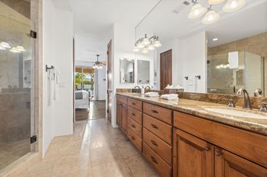 Bright master bathroom with double-sink granite vanity and wood cabinets, large mirror, glass walk-in shower and tile floor, leading to a sunlit bedroom and patio with palm trees.