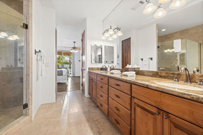 Bright master bathroom with double-sink granite vanity and wood cabinets, large mirror, glass walk-in shower and tile floor, leading to a sunlit bedroom and patio with palm trees.