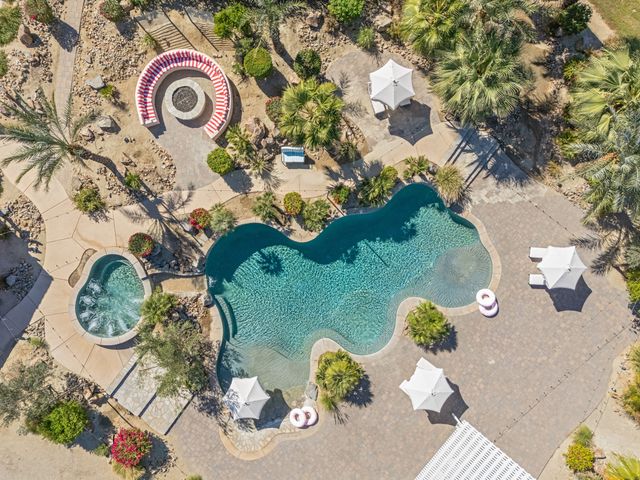 Aerial drone view of a sparkling resort-style turquoise pool and adjacent hot tub, surrounded by palm trees, white sun umbrellas, striped circular fire‑pit seating, inflatable pool rings and desert landscaping.