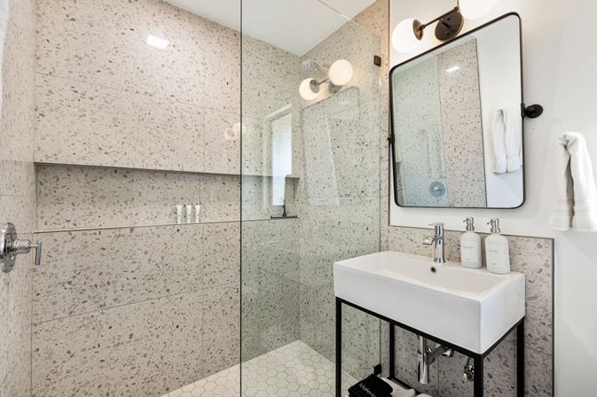 Modern residential bathroom with glass walk-in shower and terrazzo wall tiles, white hexagon floor tiles, rectangular white sink on a black metal vanity, black-framed mirror and round wall sconce lighting.