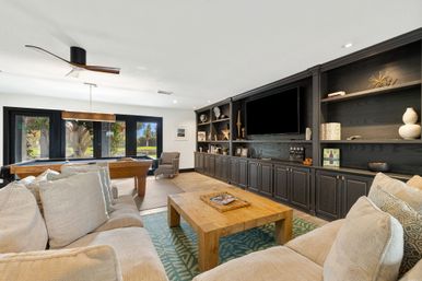 Spacious modern family game room with a beige sectional and patterned pillows, wooden coffee table, pool table by large black-framed windows overlooking a green yard, dark built-in entertainment shelves with TV, and a sleek ceiling fan.