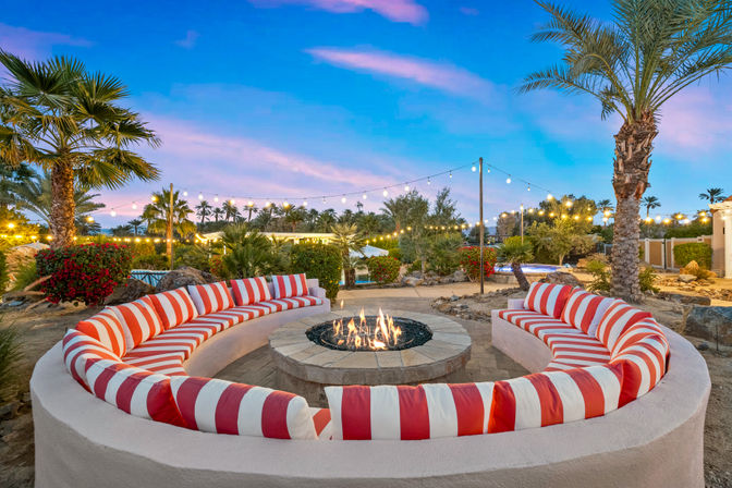 Curved outdoor fire-pit lounge with red-and-white striped cushions, twinkling string lights, palm trees and desert landscaping at sunset