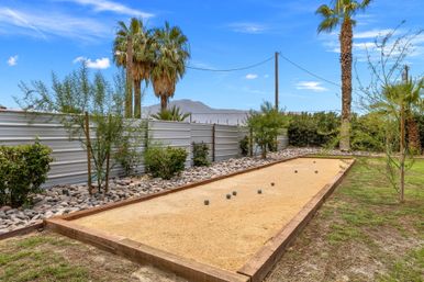 Sunny desert backyard bocce court with scattered bocce balls, framed by palm trees, river-rock beds and a corrugated metal fence, mountains and blue sky in the distance.