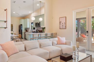 Sunny open-plan living room and pastel-green kitchen with a plush off-white bouclé sofa and blush pillows, rattan bar stools at a breakfast island, glass doors opening to a sunny patio with wicker chairs and palm trees.