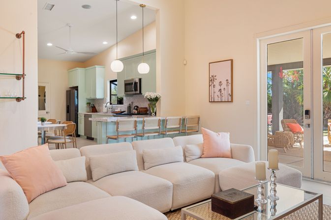 Sunny open-plan living room and pastel-green kitchen with a plush off-white bouclé sofa and blush pillows, rattan bar stools at a breakfast island, glass doors opening to a sunny patio with wicker chairs and palm trees.