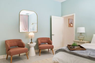 Cozy modern bedroom corner with mint-green walls, two dusty-rose mid-century chairs beside a white side table and vintage fringe lamp, arched gold-framed mirror above, nightstand with glass lamp, and bed with gray throw and wicker tray holding a small succulent.