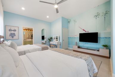 Bright coastal-themed guest bedroom with two white twin beds, light-blue walls, palm-tree and diver mural, wall-mounted TV, vanity desk, ceiling fan, and en-suite bathroom