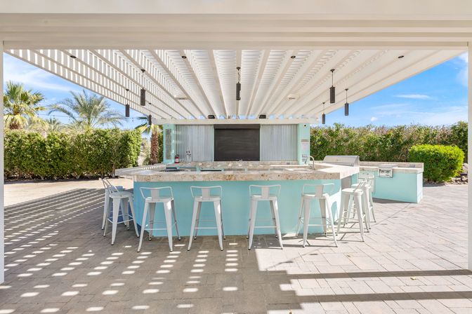 Shaded outdoor bar under a white pergola with a turquoise counter, white metal bar stools, hanging pendant lights, palm trees and sunny patio pavers