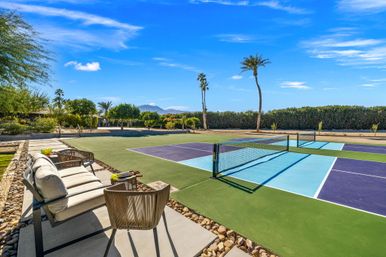 Sunny outdoor pickleball courts with blue and purple courts, net and paddles on a patio seating area, palm trees and desert landscaping with a mountain backdrop under a clear blue sky