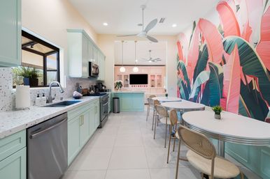 Sunlit modern kitchen with mint-green cabinets, terrazzo countertops and stainless appliances, a breakfast bar and retro oval diner tables with rattan chairs beside a large pink-and-green tropical banana-leaf mural.