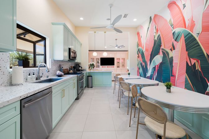 Sunlit modern kitchen with mint-green cabinets, terrazzo countertops and stainless appliances, a breakfast bar and retro oval diner tables with rattan chairs beside a large pink-and-green tropical banana-leaf mural.