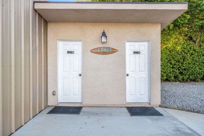 Outdoor restroom entrance with two white doors flanking a surfboard-shaped 'Baños' sign on a stucco wall, lantern above, rubber mats on a concrete pad, metal siding left and green shrubs right.