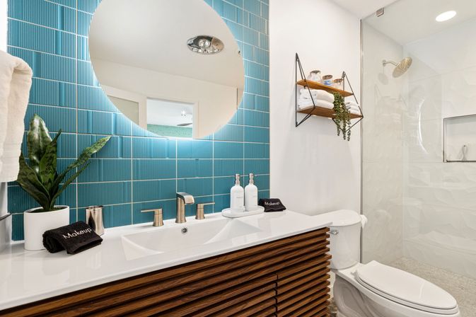 Bright modern bathroom with teal vertical tiles, round backlit mirror above a white sink and walnut slatted vanity, chrome faucet, potted plant, floating wood shelves with towels, and a glass walk-in shower.