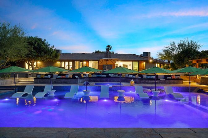 Resort-style twilight backyard pool with purple LED lights, in-water lounge chairs and green umbrellas, patio seating and modern single-story home in background.