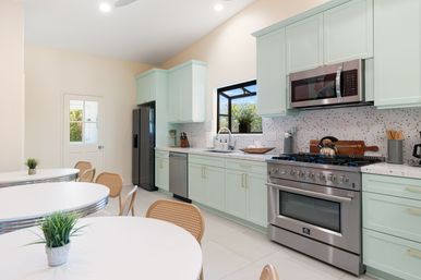 Bright modern kitchen with mint-green cabinets, terrazzo backsplash, stainless-steel range and fridge, round white diner tables with rattan chairs and a window plant