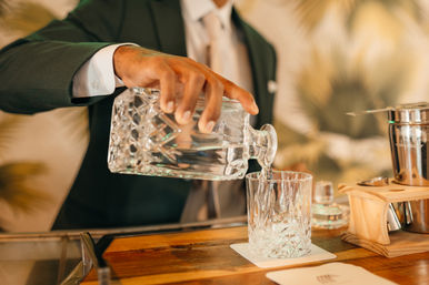 Bartender in a suit pours water from a cut-glass decanter into a crystal tumbler on a wooden bar counter, elegant cocktail lounge vibe.