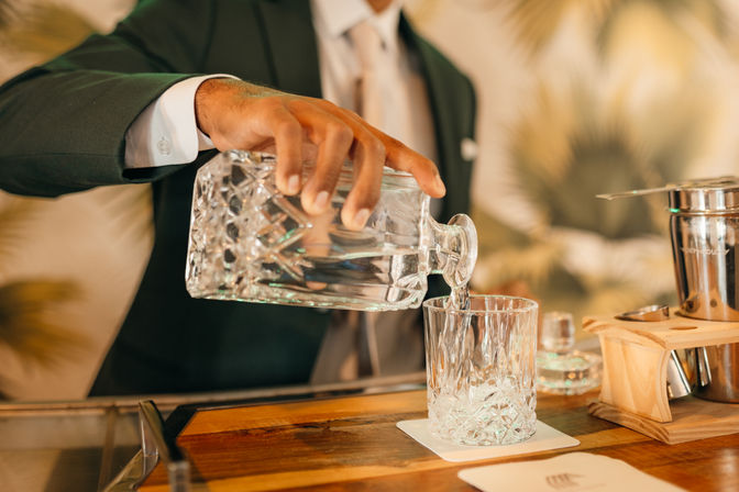 Bartender in a suit pours water from a cut-glass decanter into a crystal tumbler on a wooden bar counter, elegant cocktail lounge vibe.