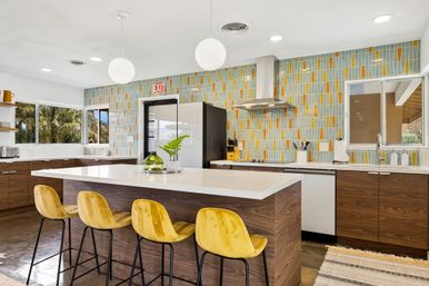 Sunny modern kitchen with a large white island, four mustard-yellow velvet bar stools, wood lower cabinets, geometric teal-and-orange tile backsplash, stainless range hood, globe pendant lights and windows showing palm trees.