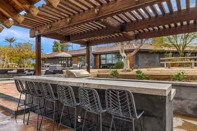 Outdoor stone bar and grill with woven bar stools under a wooden pergola, modern backyard patio with lounge seating, pool edge, and drought-tolerant landscaping