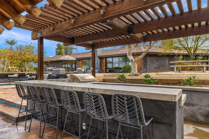 Outdoor stone bar and grill with woven bar stools under a wooden pergola, modern backyard patio with lounge seating, pool edge, and drought-tolerant landscaping