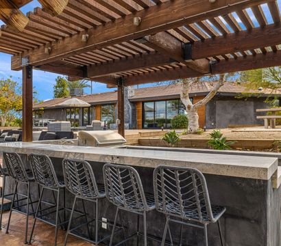 Outdoor stone bar and grill with woven bar stools under a wooden pergola, modern backyard patio with lounge seating, pool edge, and drought-tolerant landscaping